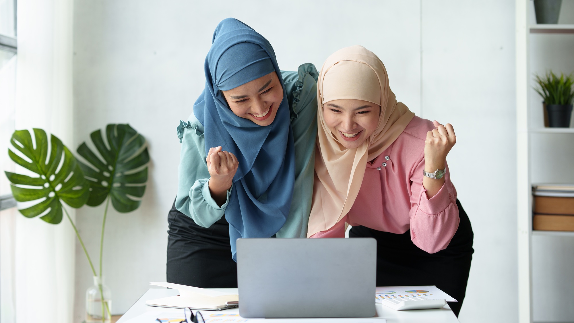 Two happy businesswomen in hijab celebrate success while working together on a laptop in an office